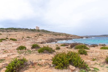 Bulutlu bir günde turkuaz Akdeniz manzaralı bir tepenin üzerinde tarihi bir taş kule bulunan engebeli kayalık bir kıyı şeridinin panoramik görüntüsü.