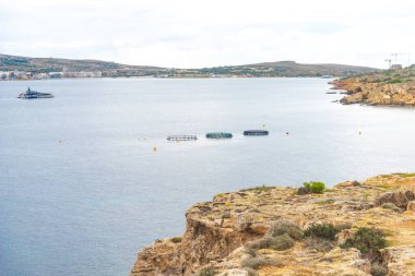 Mavi denizde yüzen üç dairesel balık çiftliği ön planda kayalık uçurumlar ve bulutlu bir gökyüzünün altındaki uzak bir sahil kasabasıyla çevrili geniş bir çekim..