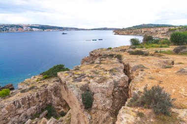 Akdeniz manzaralı bulutlu bir gökyüzünün altında uzak bir sahil kasabası ve tepeleri olan sakin mavi bir deniz koyuna bakan engebeli kireçtaşı kayalıklarından manzara.