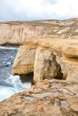 Yüksek katmanlı kumtaşı kayalıklarının dikey görüntüsü engebeli mavi denize çarpan dalgalar ve bulutlu hava altında kayalık bir kıyı şeridinde beyaz köpükle kavuşuyor..
