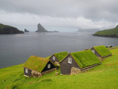 Faroe Adaları 'nın yeşil manzaraları. Tavanında yeşil çimen olan geleneksel Faroe siyah evleri, Drangarnir kaya oluşumu, Tindhomur adası ve Atlantik Okyanusu ufku. Vagar Adası.