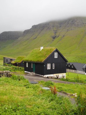 Faroe Adaları 'nın yeşil manzaraları. Çatısı yeşil çimlerle kaplı geleneksel siyah Faroe evi ve Gasadalur köyü, Vagar Adası 'ndaki bulut kaplı yamaçlar..