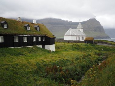 Faroe Adaları 'nın yeşil kuzey manzaraları. Beyaz kilise ve geleneksel Faroe evleri Vidareidi köyünün çatısında yeşil çimenler olan Vidoy adası fiyortlarının arasında..