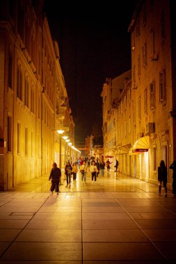 Gece şehir ışıkları, dar bir sokakta yürüyen insanlar.