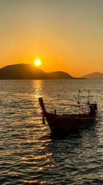 Gün batımının altın ışıklarıyla uzun kuyruklu bir tekne sakin sularda sürüklenir. Yumuşak güneş ışığı dalgaların dokusunu ve dağların siluetini vurgular. Tayland, Phuket