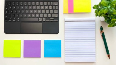 Temiz bir defter, dizüstü bilgisayar, parlak renkli yapışkan notlar ve bir bitkinin yanındaki beyaz masada bir kalem. Planlama, öğrenme, görev organizasyonu ve üretkenlik kavramları.