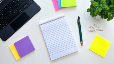 Boş bir not defteri, kalem, renkli yapışkan notlar, dizüstü bilgisayar ve beyaz bir masada ataç. Planlama, görev organizasyonu ve verimlilik kavramı.