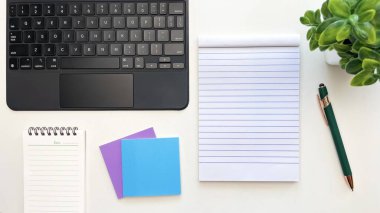 Temiz bir defter, dizüstü bilgisayar, renkli yapışkan notlar ve bir bitkinin yanındaki beyaz masada bir kalem. Planlama, ofis işi ve görev organizasyonu kavramı.
