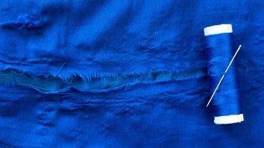 Uzun bir yırtığı olan hasarlı mavi kumaş. Yakınlarda bir makara mavi ip ve dikiş iğnesi var. Giysi tamiri ve tekstil yenileme kavramı..