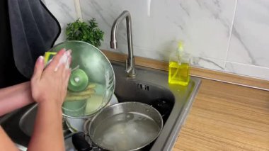Bir kadın mutfak lavabosunda tencere kapağını süngerle ve bulaşık deterjanıyla temizliyor. Temizlik ve evde bulaşık yıkama kavramı.