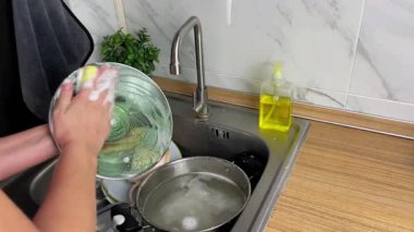 Genç bir kadın mutfakta bulaşık deterjanıyla cam tencere kapağını yıkıyor. Ev hayatı ve temizlik kavramı.