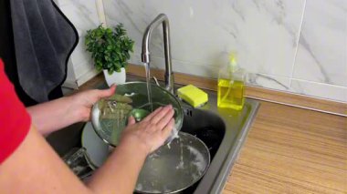 Bir kadın cam tencere kapağını dikkatlice duruluyor, akan suyun altındaki lavaboda deterjanı yıkıyor. Hijyen anlayışı ve evde temizlik sağlanması.