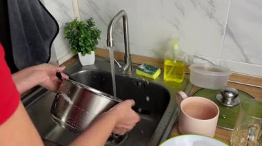 Bir kadın tencereyi elle yıkar, deterjanı temizlemek için suyun altında durulandırır. Yemek pişirdikten sonra mutfağı temizleme sahnesi..