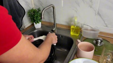 Genç bir kadın, mutfak spatulasını akan suyun altında sünger ve deterjanla yıkıyor. Ev hayatı, hijyen ve günlük mutfak temizliği kavramı..