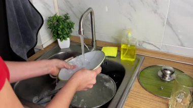 Bir kadın modern bir mutfağın lavabosunda deterjanı dikkatlice duruluyor. Yemek pişirdikten sonra düzeni ve temizliği sağlama sürecini resmeden bir ev sahnesi.