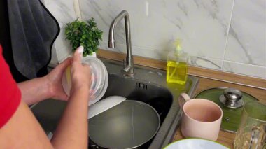 Bir kadın mutfak lavabosunda sünger ve deterjanla akan suyun altında plastik bir kabı yıkıyor. Yemek pişirdikten sonra ev hayatı, hijyen ve günlük temizlik kavramı.
