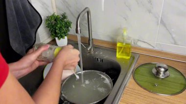 Bir kadın, mutfaktaki akan suyun altında temiz bir cam bardağı süngerle ve bulaşık deterjanıyla yıkıyor. Ev hayatı kavramı, hijyen ve bulaşıkları yıkadıktan sonra temizlik..