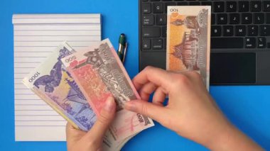 Bir dizüstü bilgisayar ve not defterinin yanında Kamboçya para birimlerini sayan el görüntüsü. Uzak çalışma kavramları, çevrimiçi kazançlar, dijital ekonomi ve gelir yönetimi.