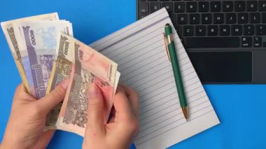 Dizüstü bilgisayarın yanında Kamboçya parası sayan eller ve mavi arka planda not defteri. İş, girişimcilik, mali işlemler ve sermaye yönetimi için kavramsal kavramlar.