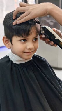 Bir kuaförde saçlarını elektrikli makasla kesen, neşeli bir tımar anı yakalayan ve modern çocukların saç stillerini taklit eden gülümseyen genç bir çocuk..