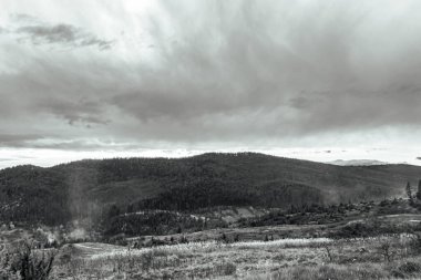 Siyah ve beyaz bulutlar bir dağ manzarası üzerinde ağaçlar ve dağlar ön planda.
