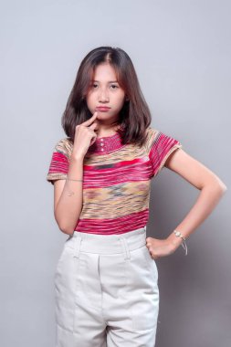 Düşünceli ve ciddi bir ifadeyle poz veren Asyalı bir kadın çenesine dokunuyor. Renkli örgü bir bluz ve yüksek bel pantolon giyiyor. Kendine güveni, tavırları ve modern gündelik tarzını gösteriyor..