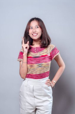 Neşeli bir Asyalı kadın, kendine güvenerek gülümserken yukarı doğru işaret ediyor, renkli çizgili bluzu ve beyaz pantolonu var. Onun parlak ifadesi yaratıcılığı, pozitifliği ve modern gençlik enerjisini aktarıyor..