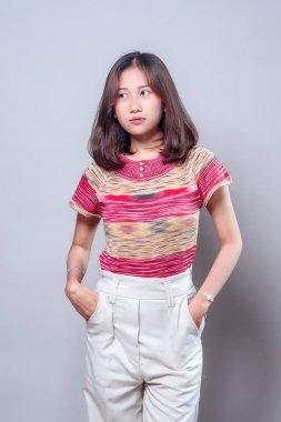 Sakin ve düşünceli bir yüz ifadesi olan Asyalı bir kadın elleri ceplerinde özgüvenle duruyor. Renkli bir bluz ve beyaz pantolon giyiyor, zarafet, tarz ve modern gündelik özgüven gösteriyor..