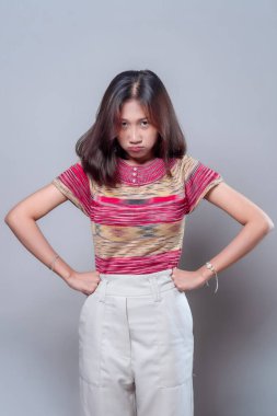 Asyalı bir kadın, elleri kalçasında, renkli çizgili bir gömlek ve açık renk pantolon giyip, güçlü duygu ve tavır sergileyen sinirli ve üzgün bir yüz ifadesi sergiliyor..