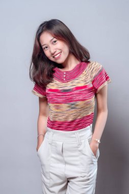 Neşeli Asyalı bir kadın şık bir kıyafet içinde, renkli çizgili bir bluz ve açık renk pantolon giyip, doğal güzelliğini ve pozitif enerjisini gösteren, kendine güvenen bir şekilde gülümsüyor..