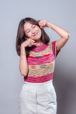 Neşeli Asyalı bir kadın sevimli bir ifadeyle, gözleri kapalı, yumrukları yüzüne yakın, renkli çizgili gömlek ve açık renk pantolon giymiş, neşe ve gençlik enerjisi gösteren bir şekilde poz veriyor..