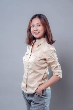 Bir eli cebinde, bej gömlek ve gri pantolon giyen, profesyonelliği, pozitifliği ve modern iş güvenini temsil eden kendine güvenen genç Asyalı bir kadın gülümsüyor..