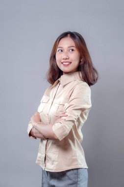 Genç Asyalı bir kadın kendinden emin bir şekilde gülümserken kollarını kavuşturmuş, bej gömlek ve gri pantolon giyiyor, iyimserliği, profesyonelliği ve modern iş güvenini simgeliyor..