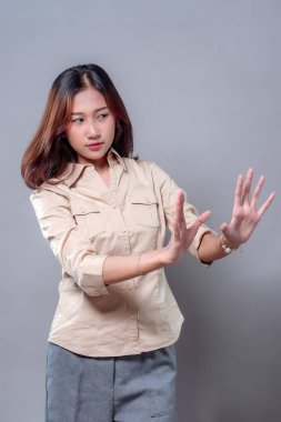 Bej gömlekli ve gri pantolonlu genç bir Asyalı kadın kendini savunmak için iki elini de kaldırıyor. Onun temkinli ifadesi ve vücut dili kibarca reddedilmeyi, kaçınmayı veya anlaşmazlığı gösteriyor..