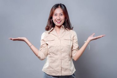 İki eli açık gülümseyen neşeli bir Asyalı kadın pozitifliğini ve güvenini dile getiriyor. Bej bir gömlek ve gri pantolon giyiyor, sıcaklığını ve profesyonelliğini gösteriyor..