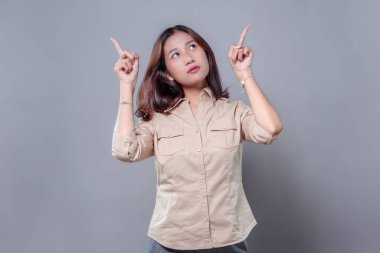 Meraklı ve düşünceli bir ifadeyle parmaklarını yukarı kaldırarak yukarı bakan genç bir Asyalı kadın. Bej bir gömlek giyiyor. İlham, ilgi ve yeni fikirleri simgeliyor..