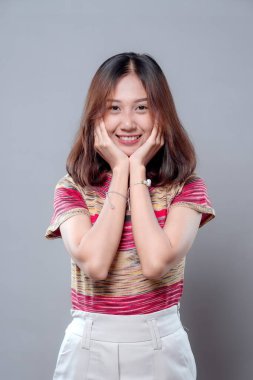 Neşeli, genç Asyalı bir kadın, elleri yanaklarında hafifçe gülümsüyor, doğal cazibesini, kendine güvenini ve pozitif duygularını modern bir kıyafetle gösteriyor..