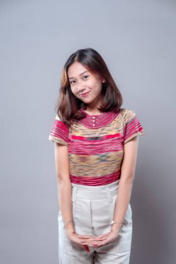 Utangaç ve nazik bir ifadeyle gülümseyen genç bir Asyalı kadın, renkli bir bluz ve beyaz pantolon giyer, özgüven, güzellik ve sıcaklığı gündelik bir görünümde gösterirken zarifçe poz verir..