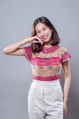 Genç Asyalı bir kadın, elleri yüzünün yanında rahatça gülümsüyor, renkli bir bluz ve beyaz pantolon giyiyor, mutluluk, sıcaklık ve pozitif enerjiyi gündelik modern tarzda ifade ediyor..