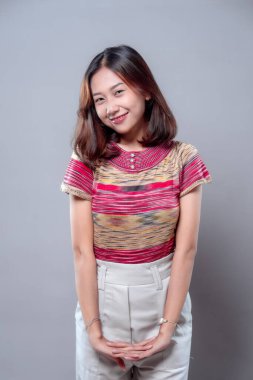 Neşeli, genç Asyalı bir kadın, elleri birarada durarak sıcak bir şekilde gülümserken, renkli bir bluz ve beyaz pantolon giyip, arkadaşlığını, zarafetini ve modern kadın cazibesini ifade ediyor..