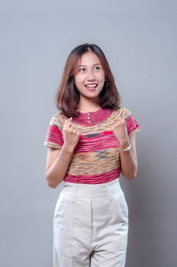 Neşeli, genç Asyalı bir kadın, heyacan dolu yumruklarıyla gülümsüyor, mutluluk, motivasyon ve iyimserliğini ifade ederken, renkli bir üst ve beyaz pantolon giyiyor, pozitif bir ruh hali içinde..