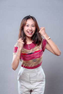 Heyecan içinde yumruklarını kaldırırken gülümseyen mutlu bir Asyalı kadın motivasyon ve başarıyı ifade ediyor. Renkli bir bluz ve beyaz pantolon giyiyor, pozitif enerji gösteriyor..