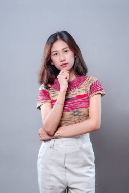 Asyalı bir kadın sakin ve düşünceli bir ifadeyle duruyor, elini çenesine koyup ileriye bakıyor. Renkli çizgili bir gömlek ve açık renk pantolon giyiyor. Kendine güvendiğini ve odaklandığını gösteriyor..