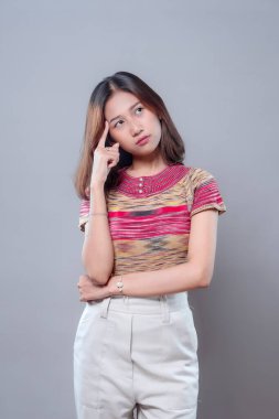 Asyalı bir kadın düşünceli bir ifadeyle duruyor, hafifçe yukarı bakarken şakağına dokunuyor. Renkli çizgili bir bluz ve bej pantolon giyiyor. Odak, zeka ve merakı temsil ediyor..
