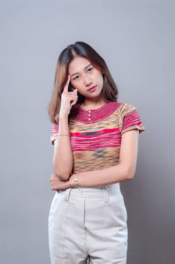 Asyalı bir kadın, bir eliyle şakağına dokunarak derin düşüncelerini ve dikkatini ifade ederek poz veriyor. Renkli çizgili bir gömlek ve açık renk pantolon giyiyor. Zeka, güven ve tefekkür taşıyor..