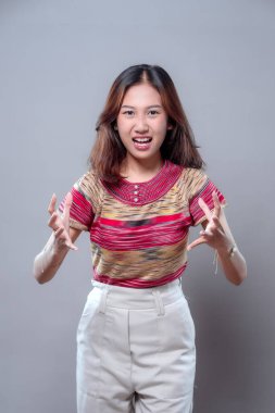 Asyalı kızgın bir kadın, elleri havada ve gergin bir yüzle hayal kırıklığını ifade ediyor. Renkli bir gömlek ve açık renk pantolon giyiyor. Güçlü duygular ve dramatik tavırlar sergiliyor..