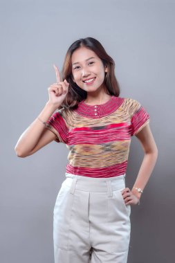 Gülümseyen Asyalı bir kadın, pozitif ve ilham verici bir tutum sergileyerek kendine güvenerek yukarıya doğru işaret ediyor. Renkli bir bluz ve açık renk pantolon giyiyor. İyimserliğini, yaratıcılığını ve modern yaşam tarzını ifade ediyor..