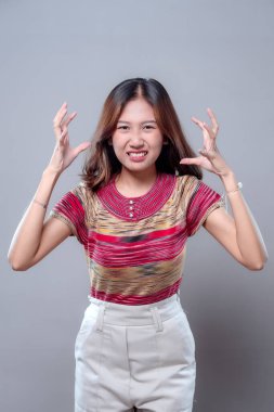 Öfkeli Asyalı bir kadın hayal kırıklığı yaşıyor, elleri havada ve gergin bir ifadeyle. Renkli çizgili bir bluz ve açık renk pantolon giyer. Günlük hayatta stres, öfke ve duygusal tepkileri tasvir eder..