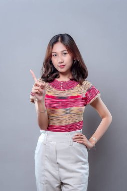 Renkli bluz ve beyaz pantolon giyen Asyalı bir kadın ciddi, kendinden emin bir ifadeyle bir parmağını kaldırır. Uyarı, hatırlatma ya da iletişimde önemli bir noktaya parmak basar..