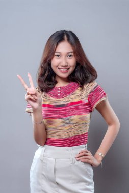 Neşeli Asyalı bir kadın parmaklarıyla zafer işareti yaparken gülümsüyor, renkli bir bluz ve açık renk pantolon giyiyor, pozitifliğini, özgüvenini ve gençlik enerjisini ifade ediyor..